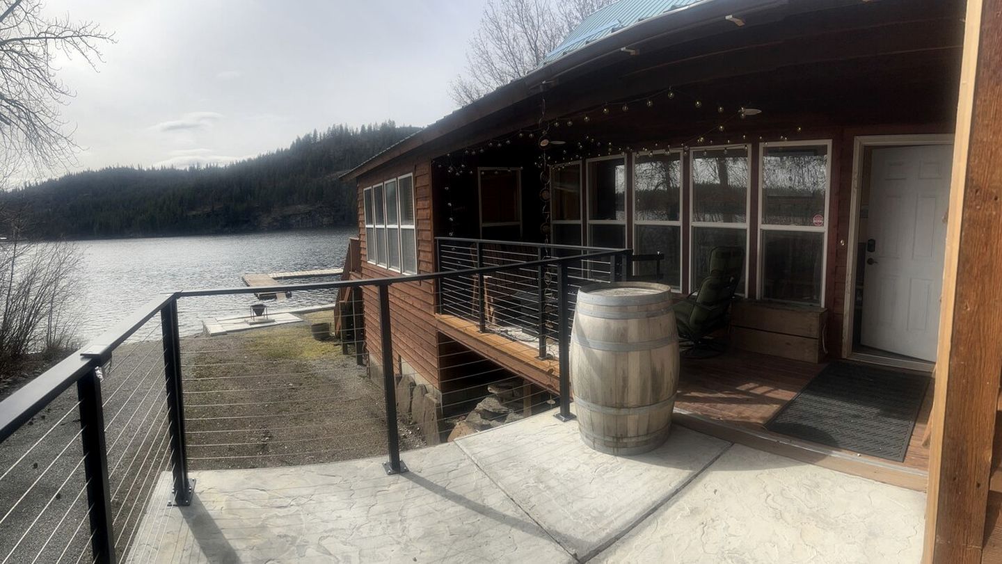 A wooden cabin with a deck overlooking a lake, a wooden barrel, and a metal railing.
