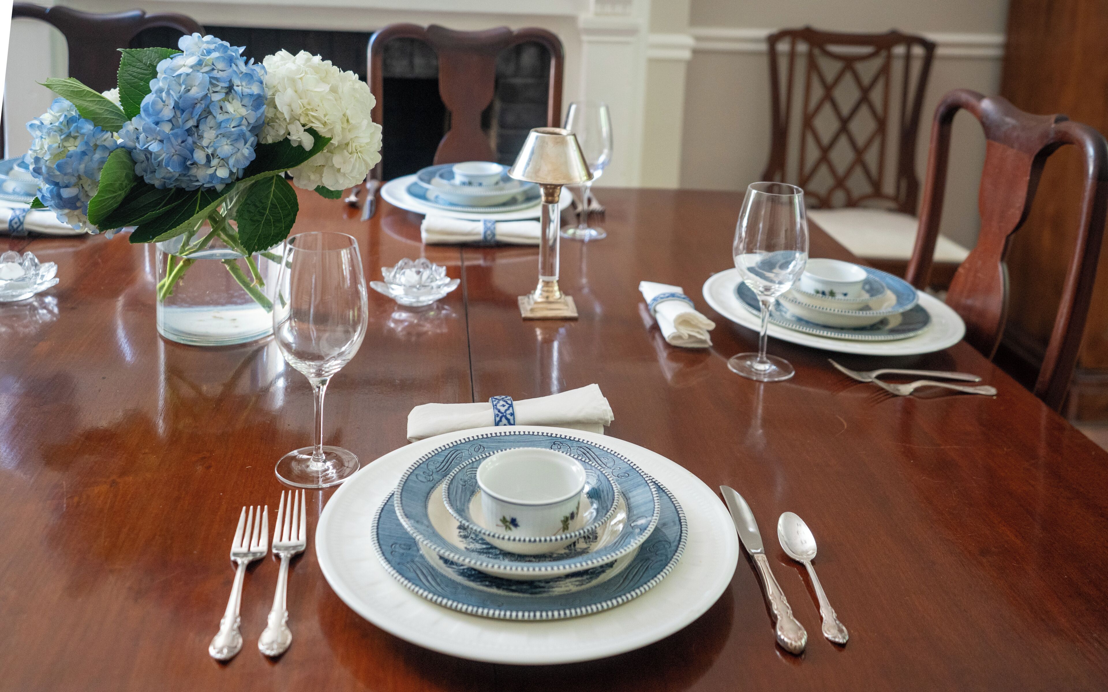 Una mesa de comedor dispuesta con platos y cubiertos azules y blancos, y un florero con flores azules y blancas.