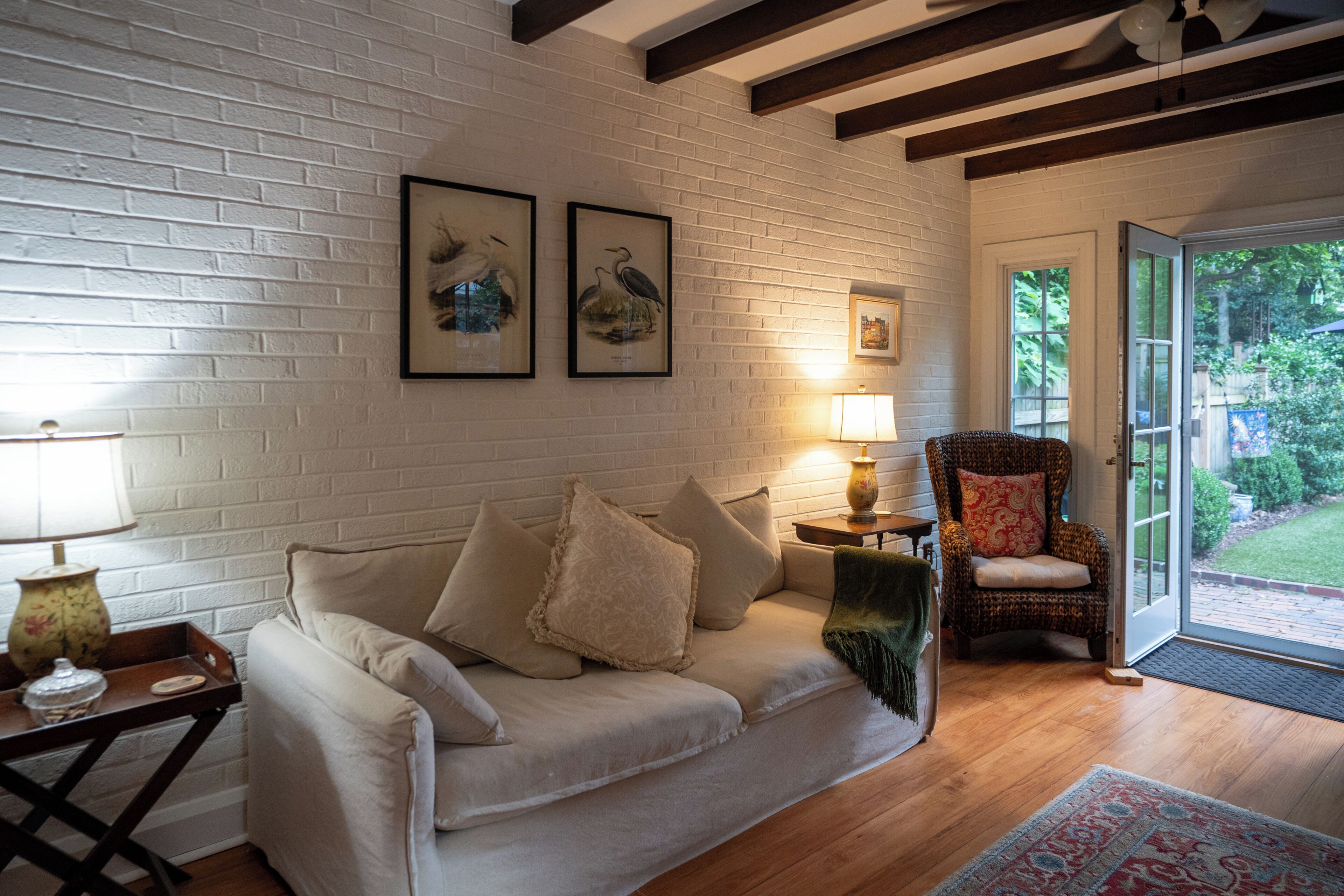 Un salón con pared de ladrillo blanco, un sofá con cojines, una silla, una lámpara y cuadros enmarcados.