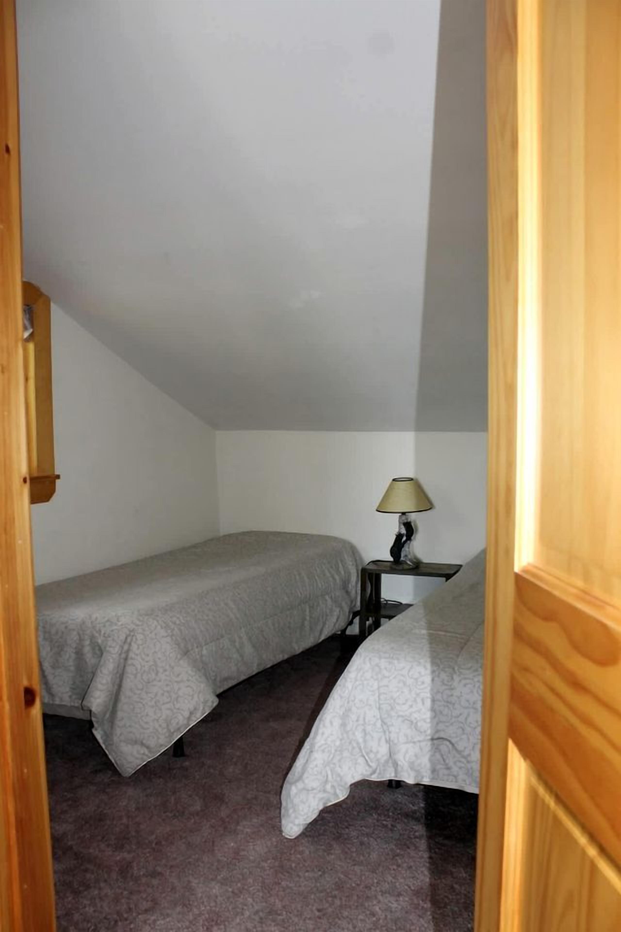 A small, single-room bedroom with two beds, a nightstand with a lamp, and a wooden door.