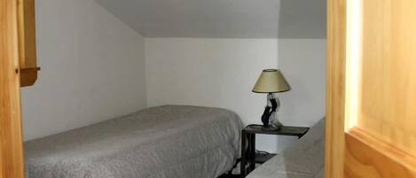 A small, single-room bedroom with two beds, a nightstand with a lamp, and a wooden door.