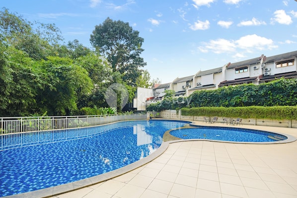Una piscina con borde de baldosas, rodeada de vegetación y un edificio con numerosas ventanas.
