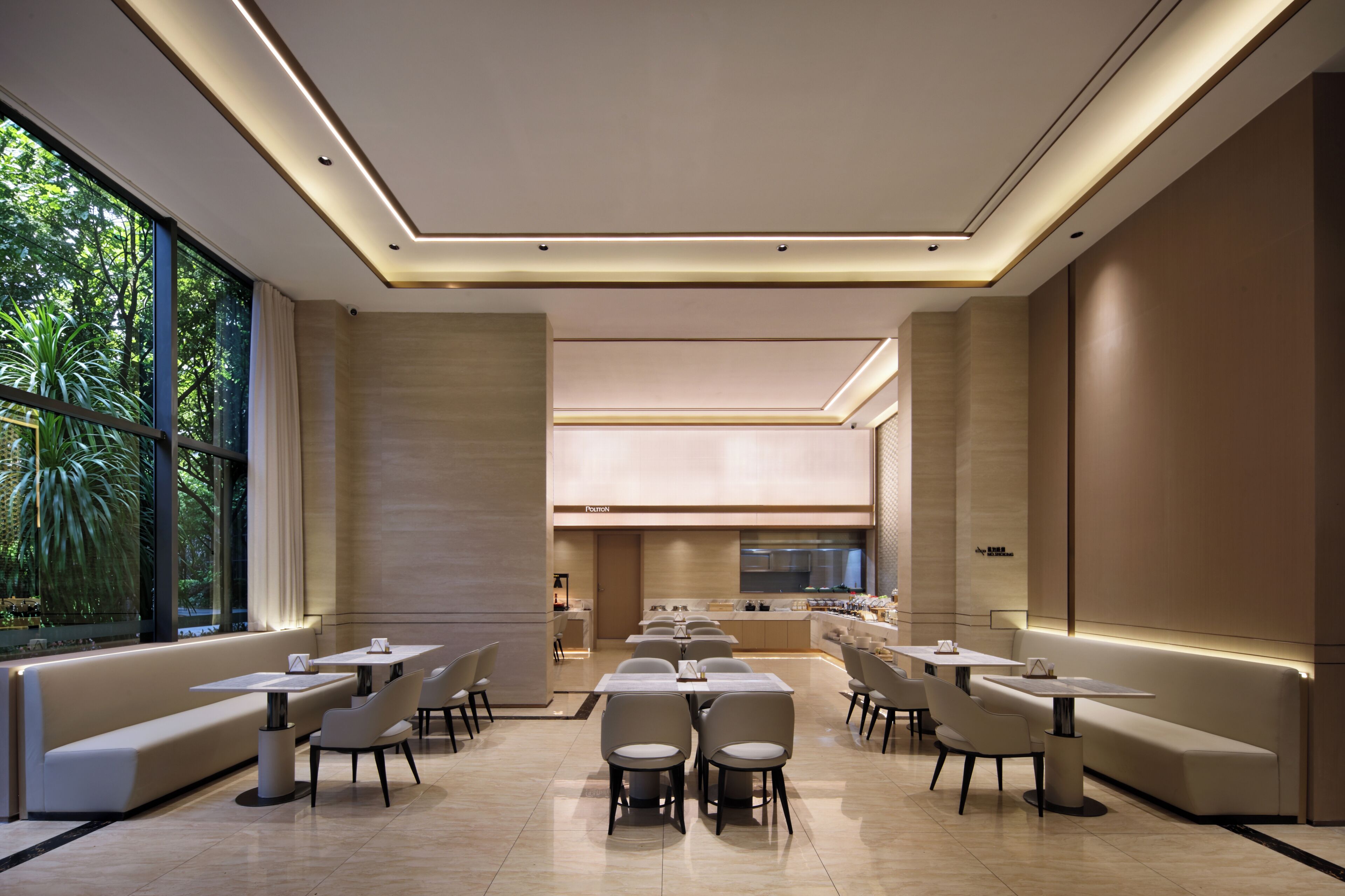 Un espace repas moderne avec un long banc, plusieurs tables et chaises, une cuisine en arrière-plan et de grandes fenêtres donnant sur un paysage verdoyant.