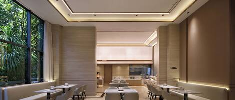 Un espace repas moderne avec un long banc, plusieurs tables et chaises, une cuisine en arrière-plan et de grandes fenêtres donnant sur un paysage verdoyant.