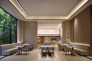 A modern dining area with a long bench, multiple tables, and chairs, a kitchen in the background, and large windows with a view of greenery.