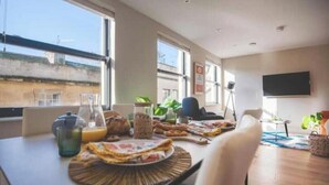Un comedor con una mesa dispuesta para el desayuno, un televisor montado en la pared y ventanas grandes que permiten la entrada de luz natural.