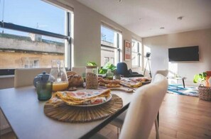 Ein Essbereich mit einem für das Frühstück gedeckten Tisch, einem an der Wand montierten Fernseher und großen Fenstern, die für natürliches Licht sorgen.