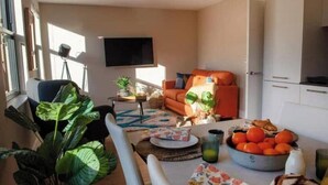 Una sala de estar con un sofá, una mesa de comedor con un cuenco de fruta y un televisor en la pared.