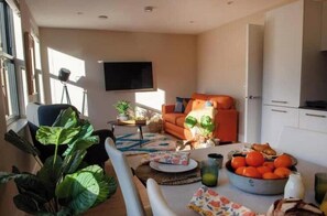 Ein Wohnzimmer mit einer Couch, einem Esstisch mit Obstschale und einem an der Wand befestigten Fernseher.