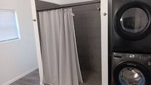 Uma lavanderia com máquina de lavar, chuveiro com cortina e janela com persianas.