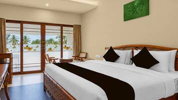 A hotel room with a large bed, a desk, chairs, and a view of palm trees through the glass doors.