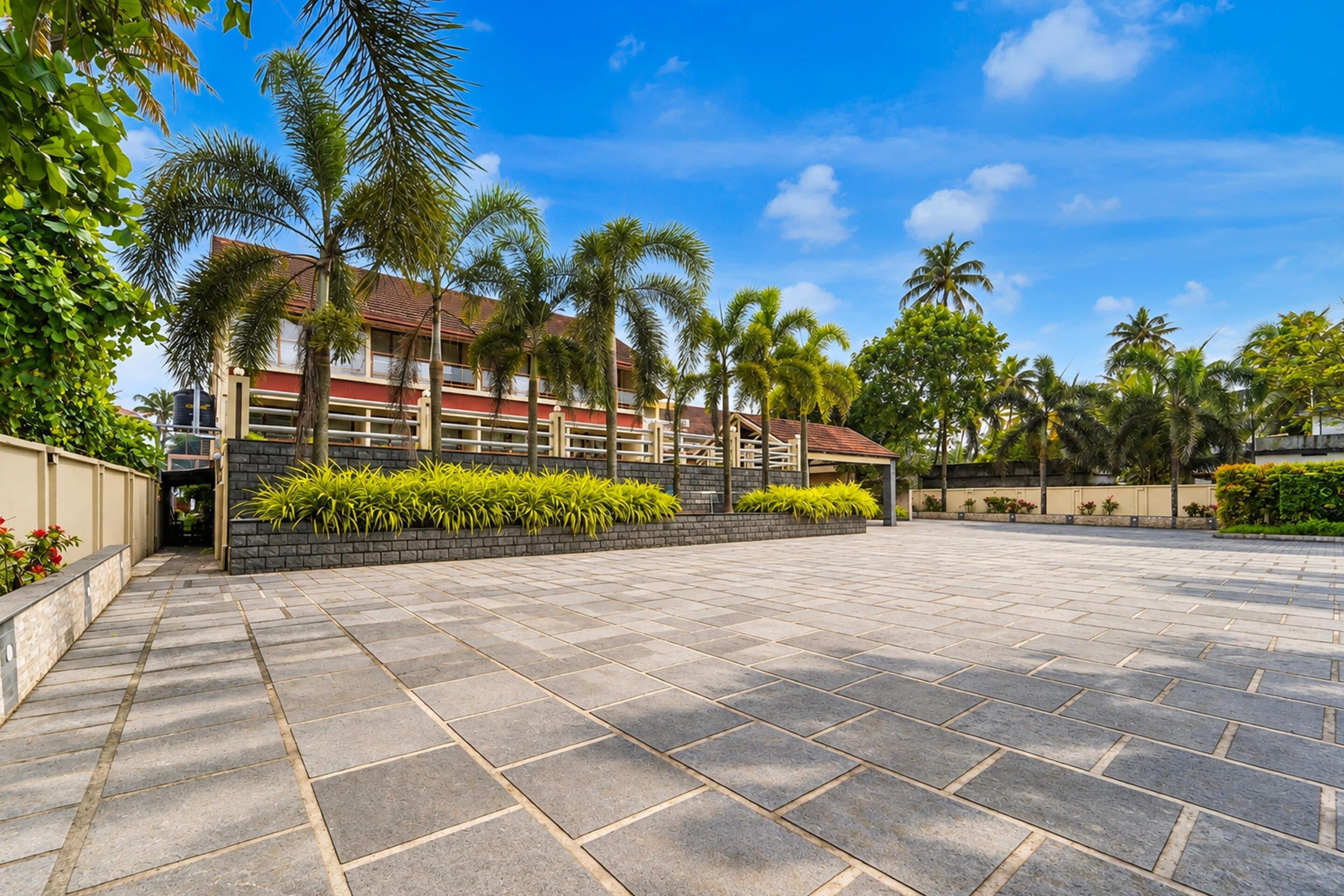 A spacious outdoor area with a paved walkway, palm trees, and a building with a red roof.
