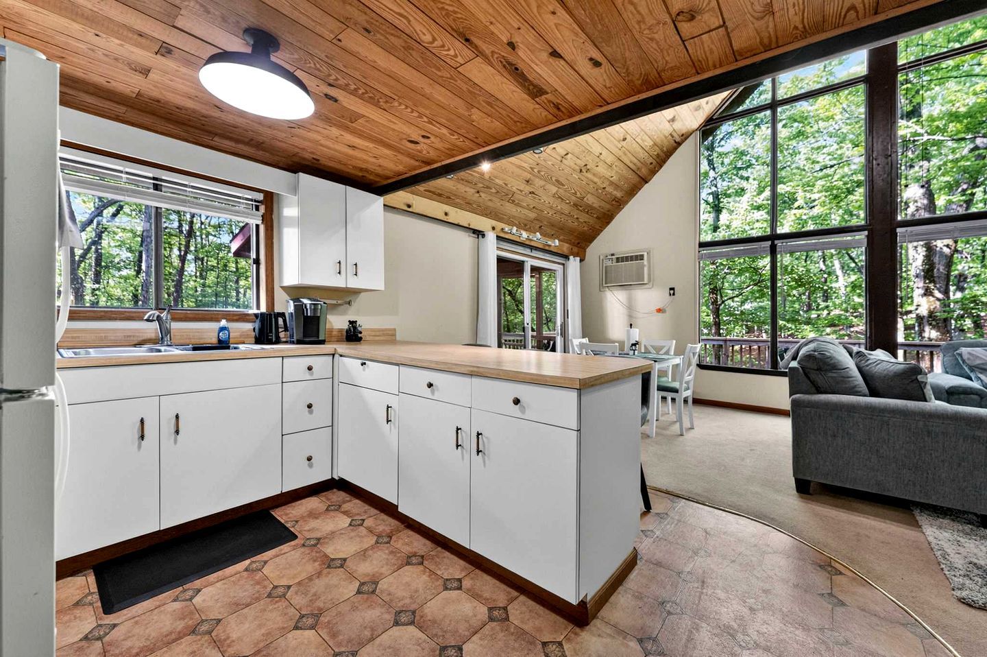 Une cuisine moderne avec des armoires blanches, un plafond en bois et de grandes fenêtres donnant sur la forêt.