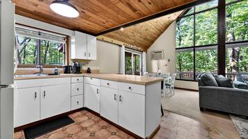 Une cuisine moderne avec des armoires blanches, un plafond en bois et de grandes fenêtres donnant sur la forêt.