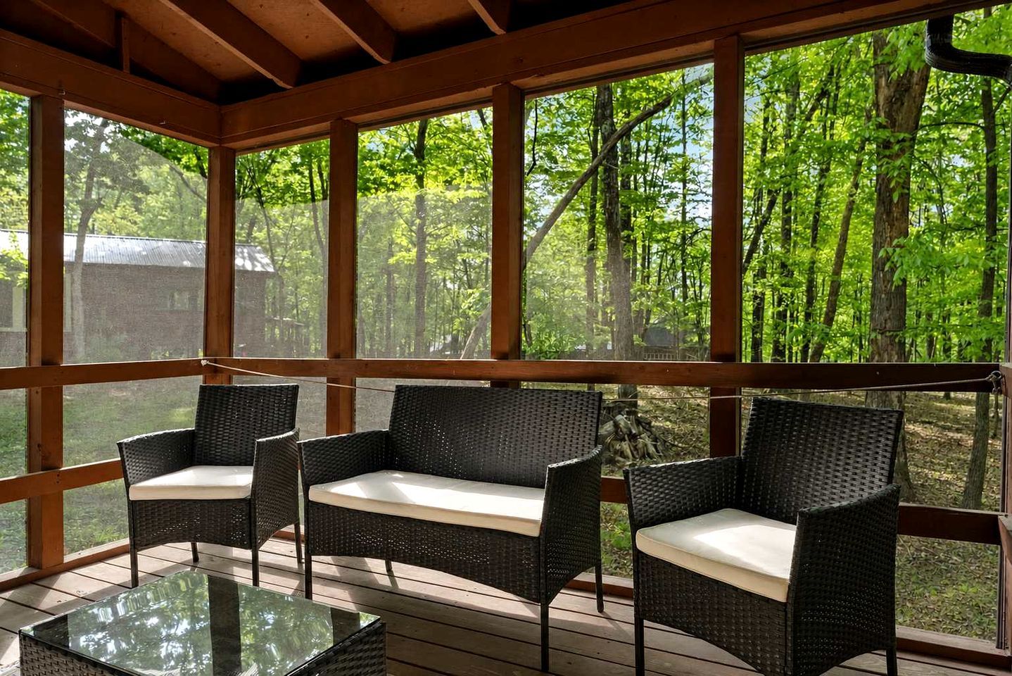 A covered outdoor seating area with wicker furniture and a glass table, overlooking a forested area.