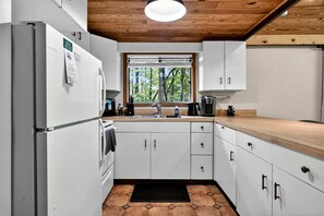 A modern kitchen with white cabinetry, a wooden countertop, a built-in oven, a sink, and a window with a view of trees.