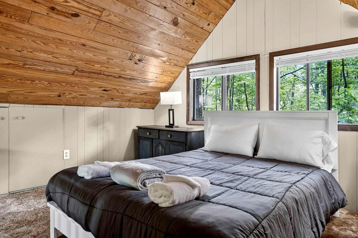 A bedroom with a wooden ceiling, a bed with a dark quilt, two bedside tables, and a window with a view of trees.