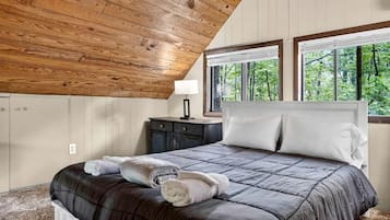 A bedroom with a wooden ceiling, a bed with a dark quilt, two bedside tables, and a window with a view of trees.