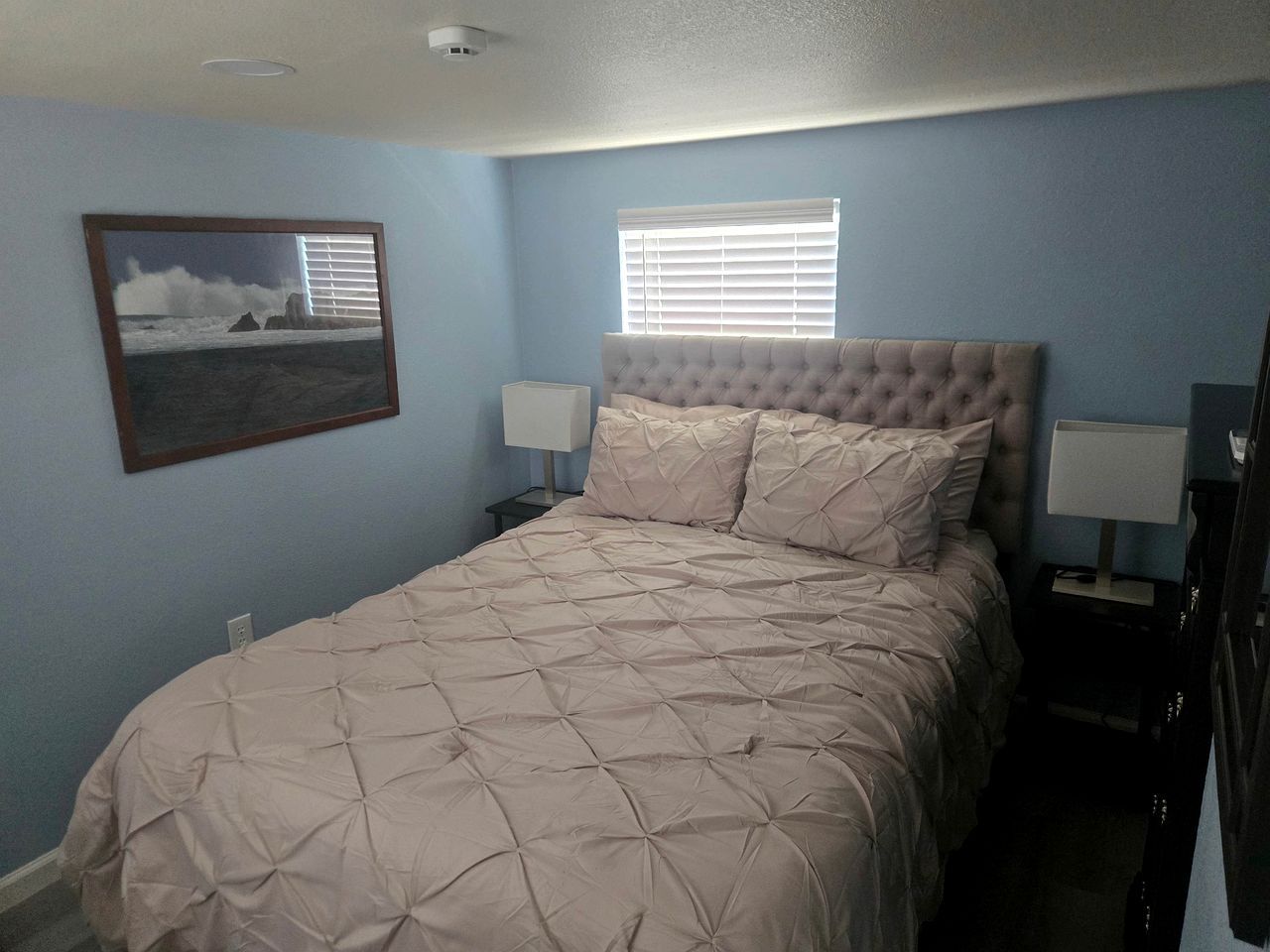 Un dormitorio con una cama, dos lámparas de noche, un ventanal con persianas y un cuadro en la pared.