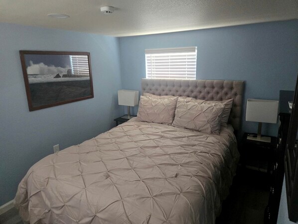 A bedroom with a bed, two bedside lamps, a window with blinds, and a framed picture on the wall.