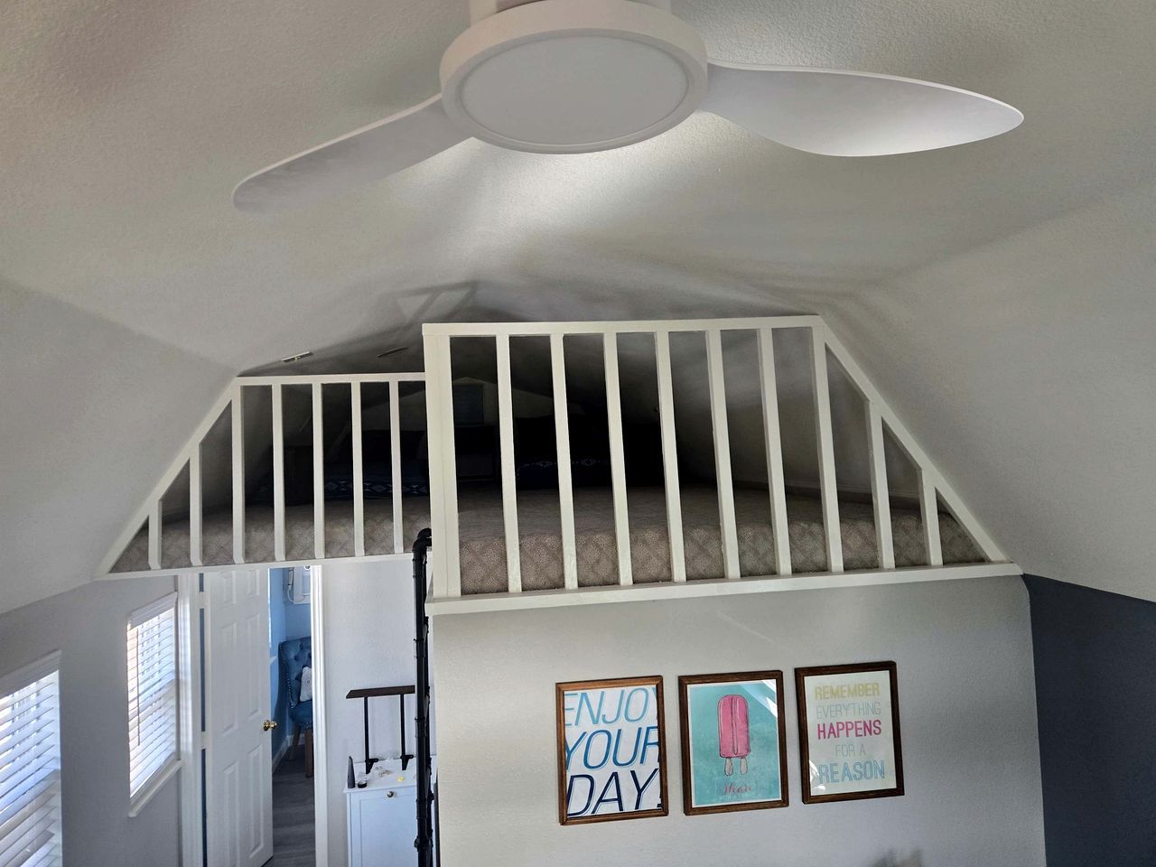 A room with a sloped ceiling, a white ceiling fan, a staircase with white railings, and three framed motivational posters on the wall.