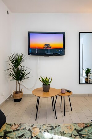 A modern living room with a flat-screen TV mounted on the wall, a round wooden table, two potted plants, a small side table with a remote control, and a patterned rug. - Aroma Holiday House with Wi-Fi and Air Conditioning (Rome)