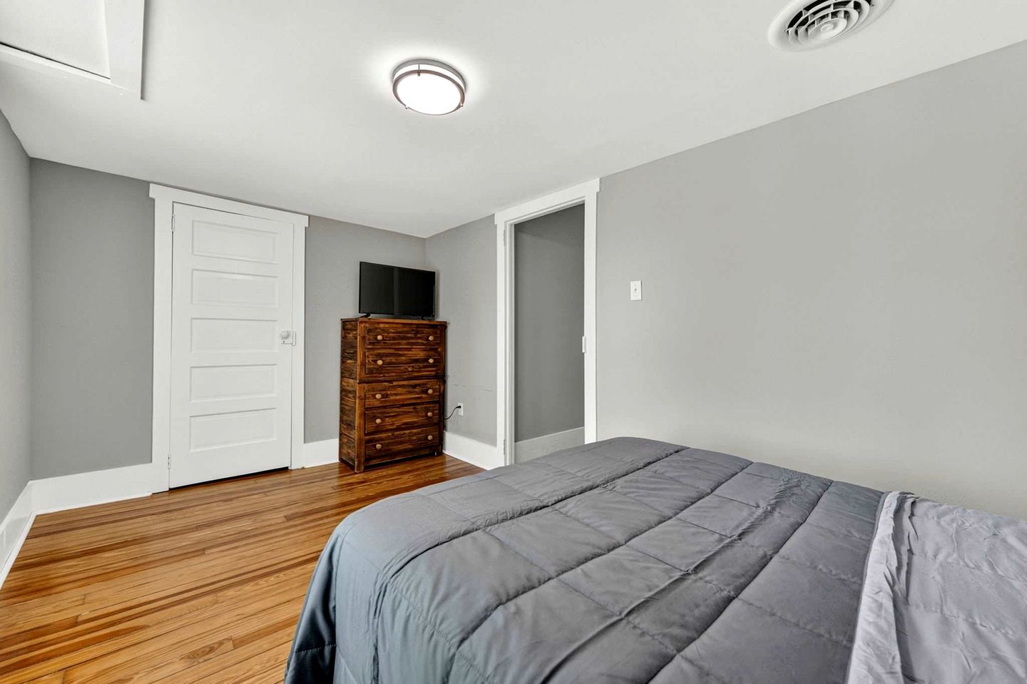 Une chambre avec une commode en bois, une couvre-lit grise et une porte blanche.