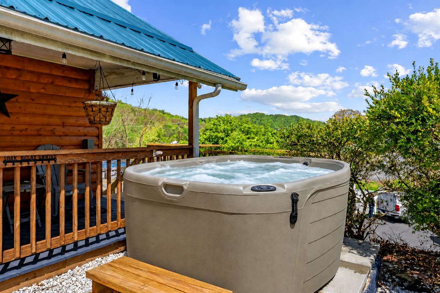 Un bain à remous à l’extérieur d’un chalet en bois doté d’un toit bleu, entouré de verdure et sous un ciel dégagé.