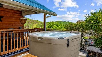 Un bain à remous à l’extérieur d’un chalet en bois doté d’un toit bleu, entouré de verdure et sous un ciel dégagé.
