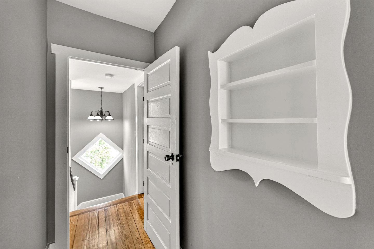 A hallway with a wooden floor, a white door, a wall-mounted shelf, and a ceiling light fixture.