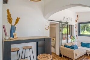 A modern living space with a kitchen area, a seating area with a sofa and cushions, and a window with a view of greenery. - Pich Home Serenity – Nature, Calm, and Genuine Connections (Miramas)