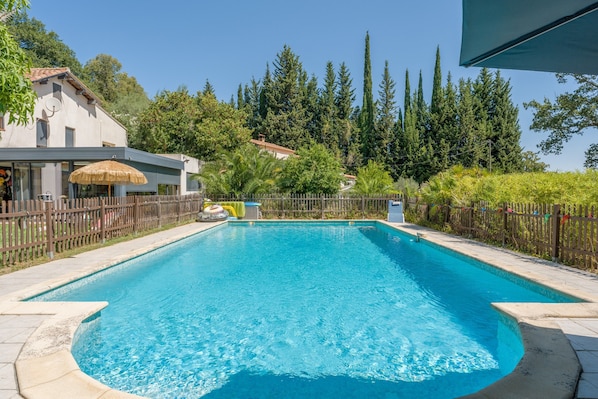 A rectangular swimming pool with a tiled edge, surrounded by a wooden fence, and a building with a satellite dish in the background.