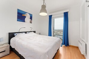 Ein Hotelzimmer mit Bett, Nachttisch, Lampe, einem Gemälde einer Möwe, einem Fenster mit blauen Vorhängen und Blick auf die Stadt.