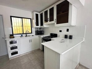 A kitchen with white cabinets, a stove, a sink, and a window with blinds. - Downtown Apartment (Santo Domingo)