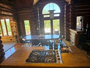 Interior de una cabaña de troncos con estufa central tipo isla, mesa de comedor y sillas.