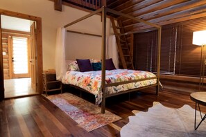 A bedroom with a four-poster bed, wooden flooring, a small bedside table, a nightstand, a window with blinds, and a lamp on a side table. - Luxury Montana-Style Log Estate with Private Vineyard & Spa Near Purcellville, VA (Purcellville)
