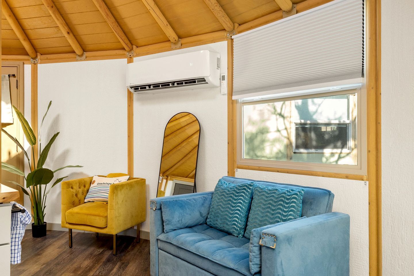 A room with a yellow armchair, a blue sofa, a wooden ceiling, an air conditioning unit, a window with blinds, and a mirror on the wall.