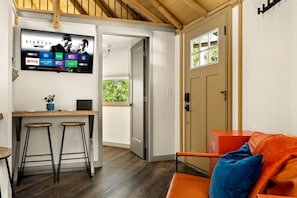 Ein kompakter Wohnbereich mit an der Wand montiertem Fernseher, der Streaming-Dienste anzeigt, einer kleinen Küchenzeile mit Barhocker und einer Tür zu einem Außenbereich.