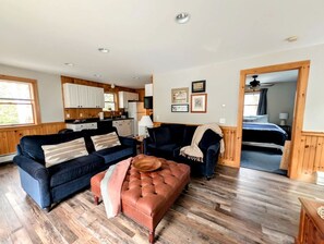 Ein großzügiger Wohnbereich mit einem dunkelblauen Sofa, einem braunen Hocker und einem Couchtisch aus Holz. Im Hintergrund ist eine Küche mit Holzausbau und Arbeitsplatte zu sehen.