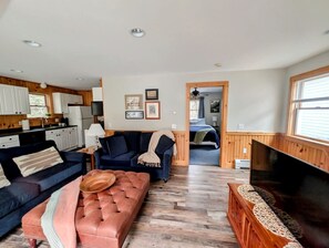 A spacious living area with a sofa, ottoman, and a flat-screen TV. A kitchen is visible in the background. - Peaceful Pet-Friendly Retreat with Workspace Near Acadia National Park in Trenton, Maine (Trenton)