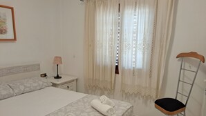 Un letto rifatto con due asciugamani arrotolati, una lampada da comodino e una sedia con seduta in legno e struttura metallica.
