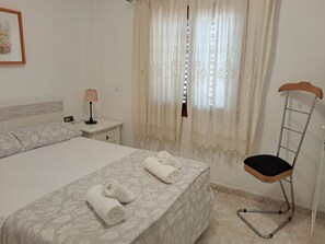 A neatly made bed with two rolled towels, a bedside lamp, and a chair with a wooden seat and metal frame.