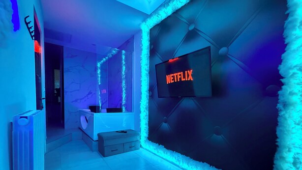 Een kamer met een televisie waarop 'NETFLIX' in rode letters staat, een witte bonttapijt en een wand met een geometrisch patroon.