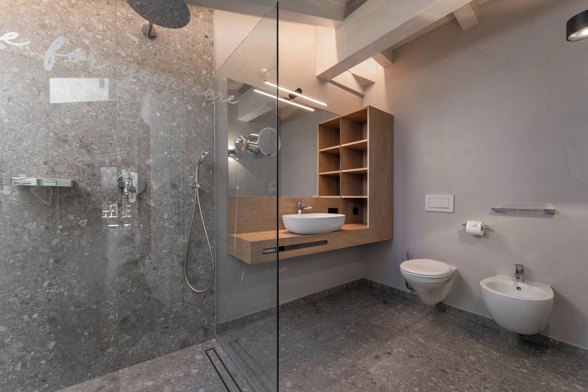 Ein modernes Badezimmer mit einer gläsernen Duschkabine, einem hölzernen Waschtisch mit Aufsatzbecken, einer Toilette und einem Bidet.