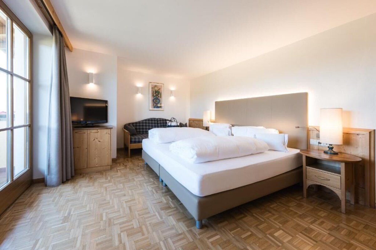 A hotel room with a large bed, a television, a desk with a lamp, and a window with curtains.