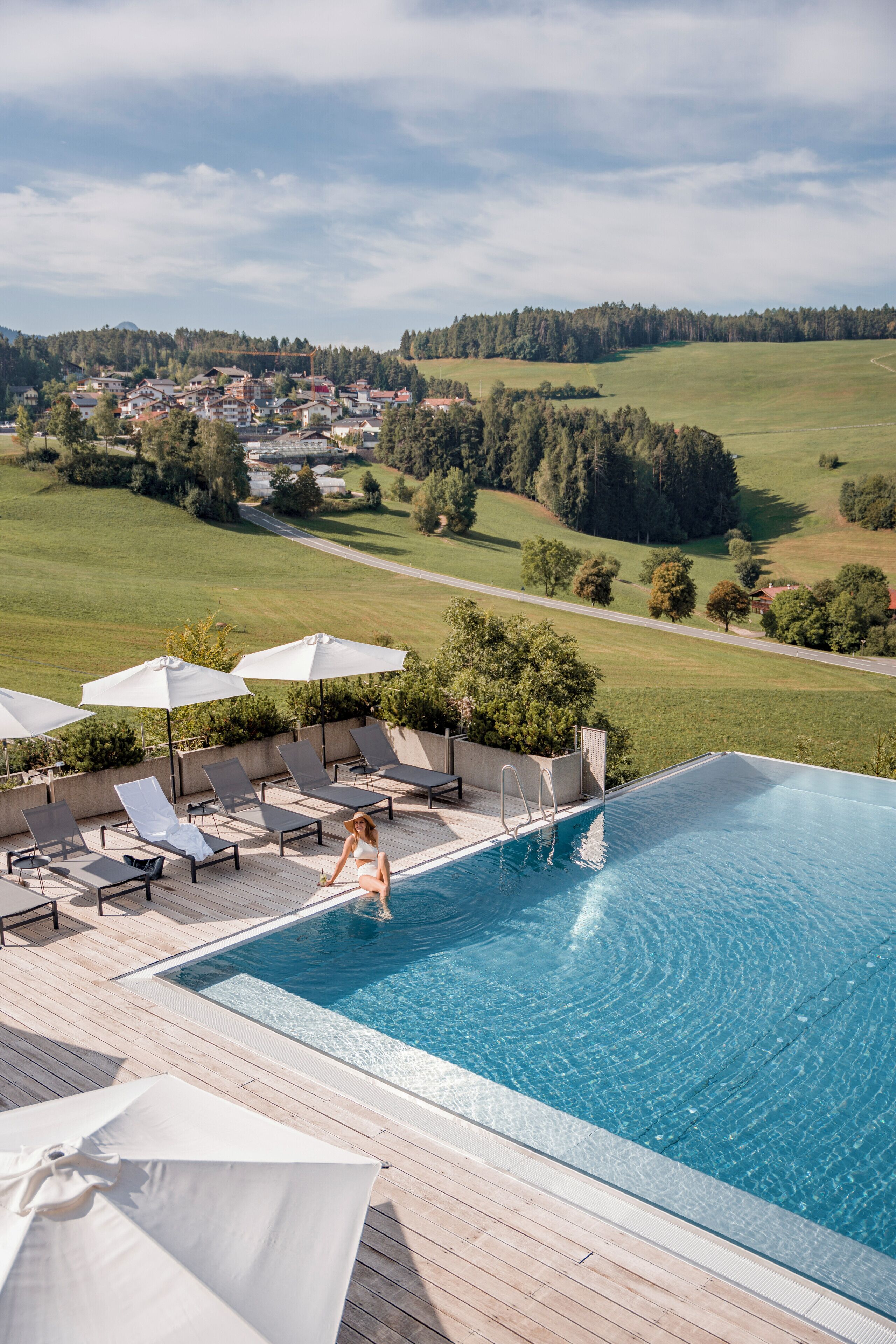 Une piscine entourée de verdure, avec des chaises longues et des parasols, d’où l’on peut admirer une vue sur une ville au loin.