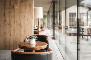 A modern outdoor seating area with a wooden table and chairs, a potted plant, and a glass wall offering a view of the interior. - Hotel Alpenflora (Castelrotto)