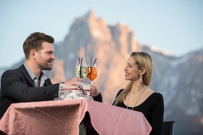 A couple toasting with drinks at a table with a mountain view in the background. - Hotel Alpenflora (Castelrotto)