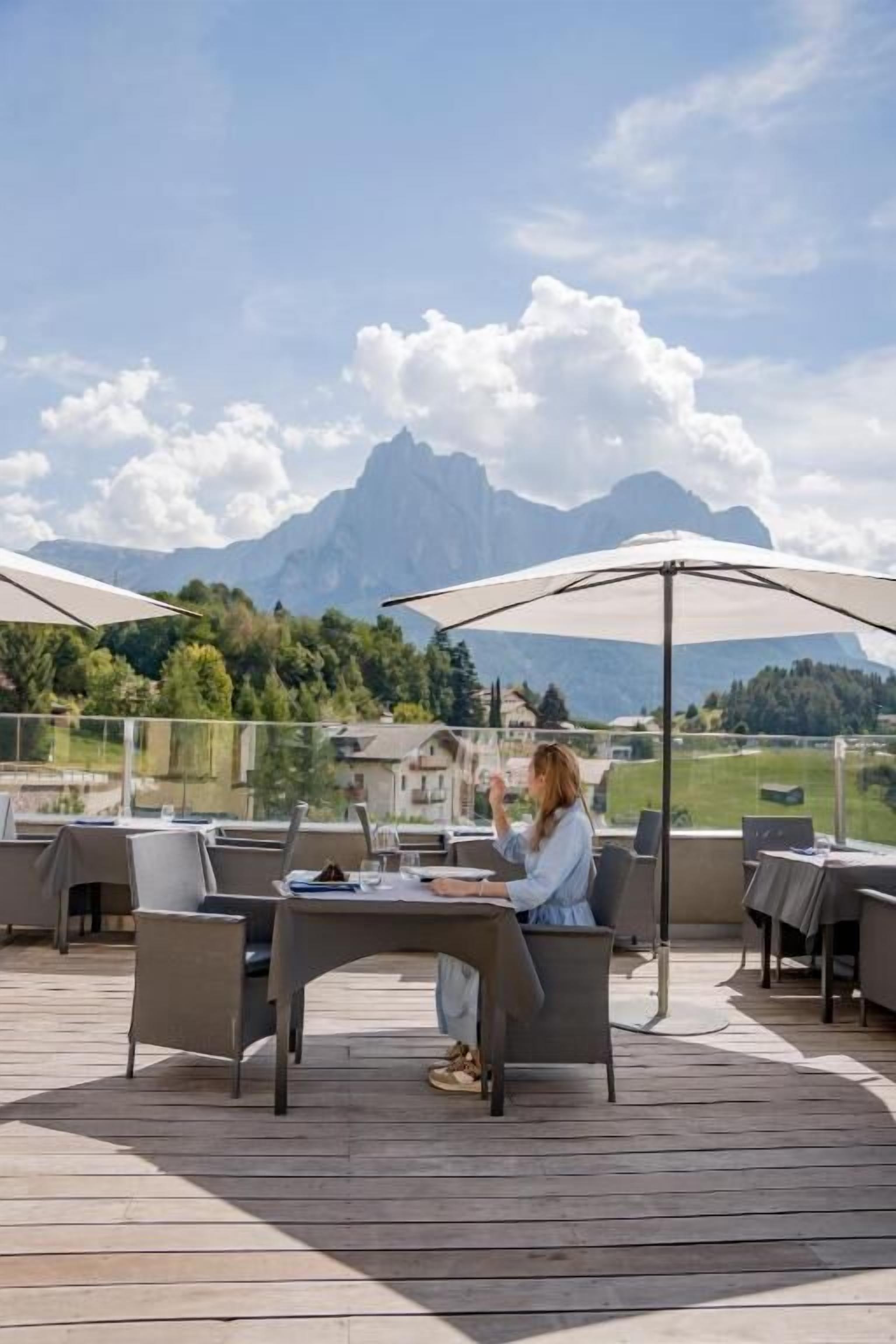 An outdoor seating area with tables and chairs, a person seated at a table, and a mountainous backdrop.