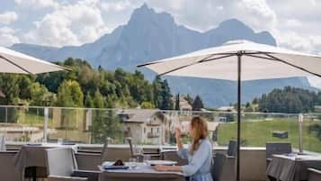 An outdoor seating area with tables and chairs, a person seated at a table, and a mountainous backdrop.